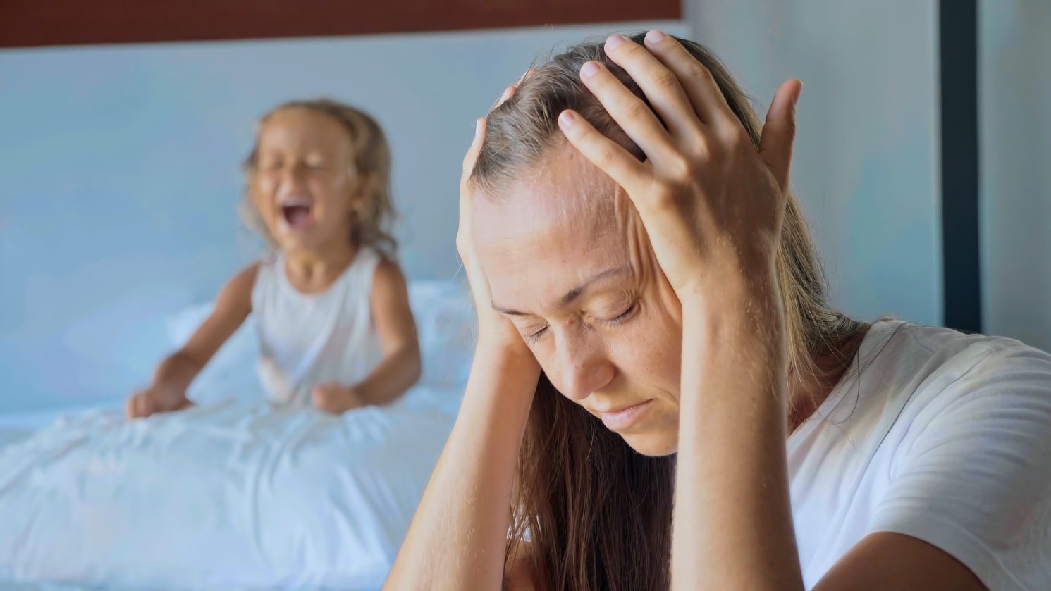 blonde hair millennial mum hands on head given up child screaming background on bed